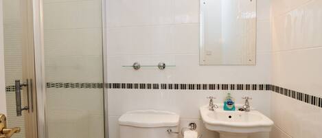 A bathroom with a glass shower enclosure, a toilet, a sink with a mirror above it, and a small shelf with toiletries.