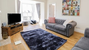 A living room with a grey sofa, a black rug, a TV on a wooden stand, and a wall art collage. - Chapel Rest (Windermere)
