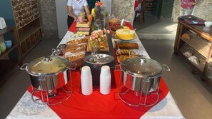 A buffet table with various food items and beverages, a person serving, and a red tablecloth. - Kontiki Hotel (São Francisco do Sul)