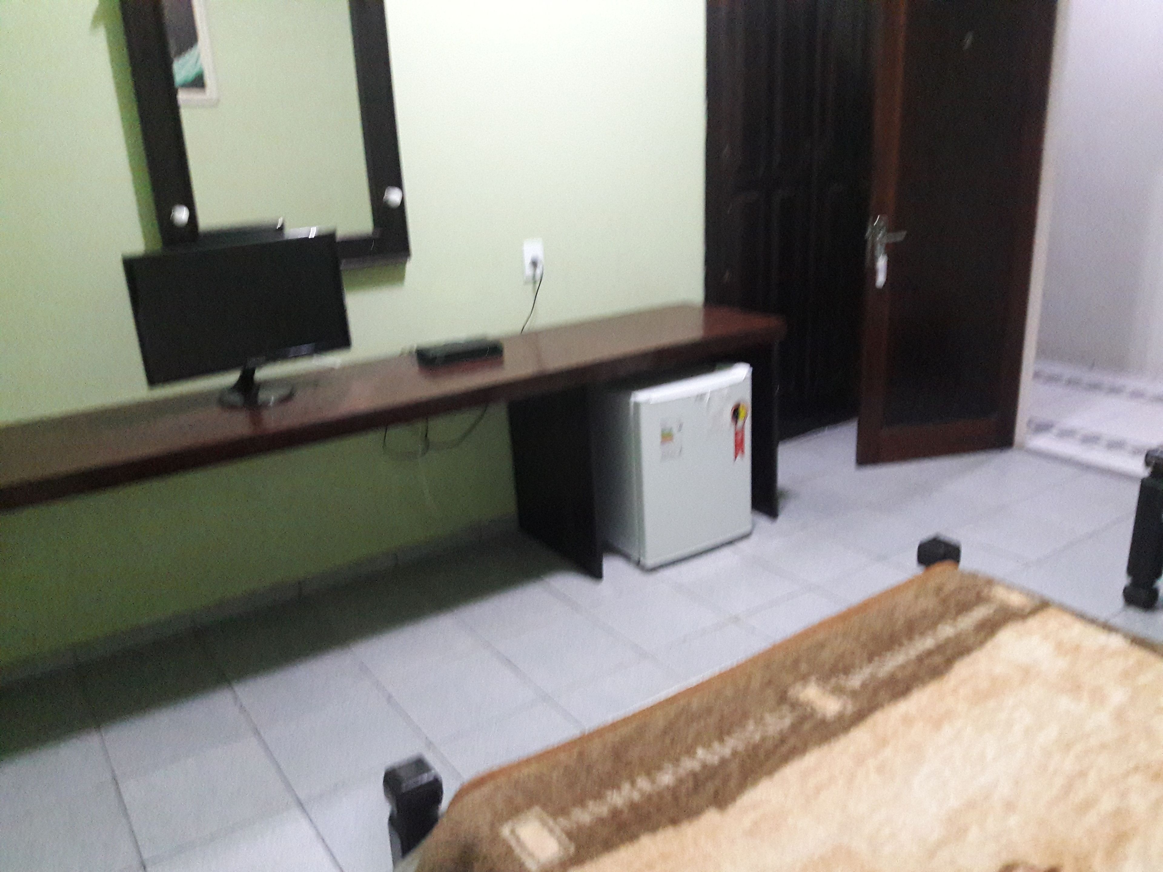 A room with a bed, a desk with a computer monitor, a small refrigerator, and a door.