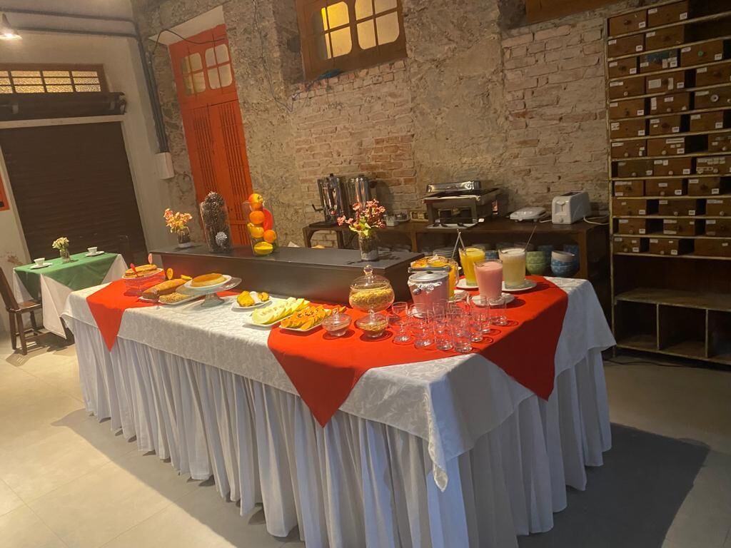 Une table de buffet garnie de divers plats et boissons, placée devant un mur de briques avec une fenêtre et une étagère remplie de boîtes.