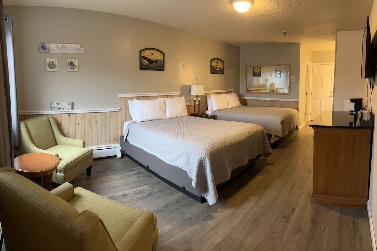 A hotel room with two beds, a sitting area with a chair and a small table, a wooden floor, and a mirror on the wall.