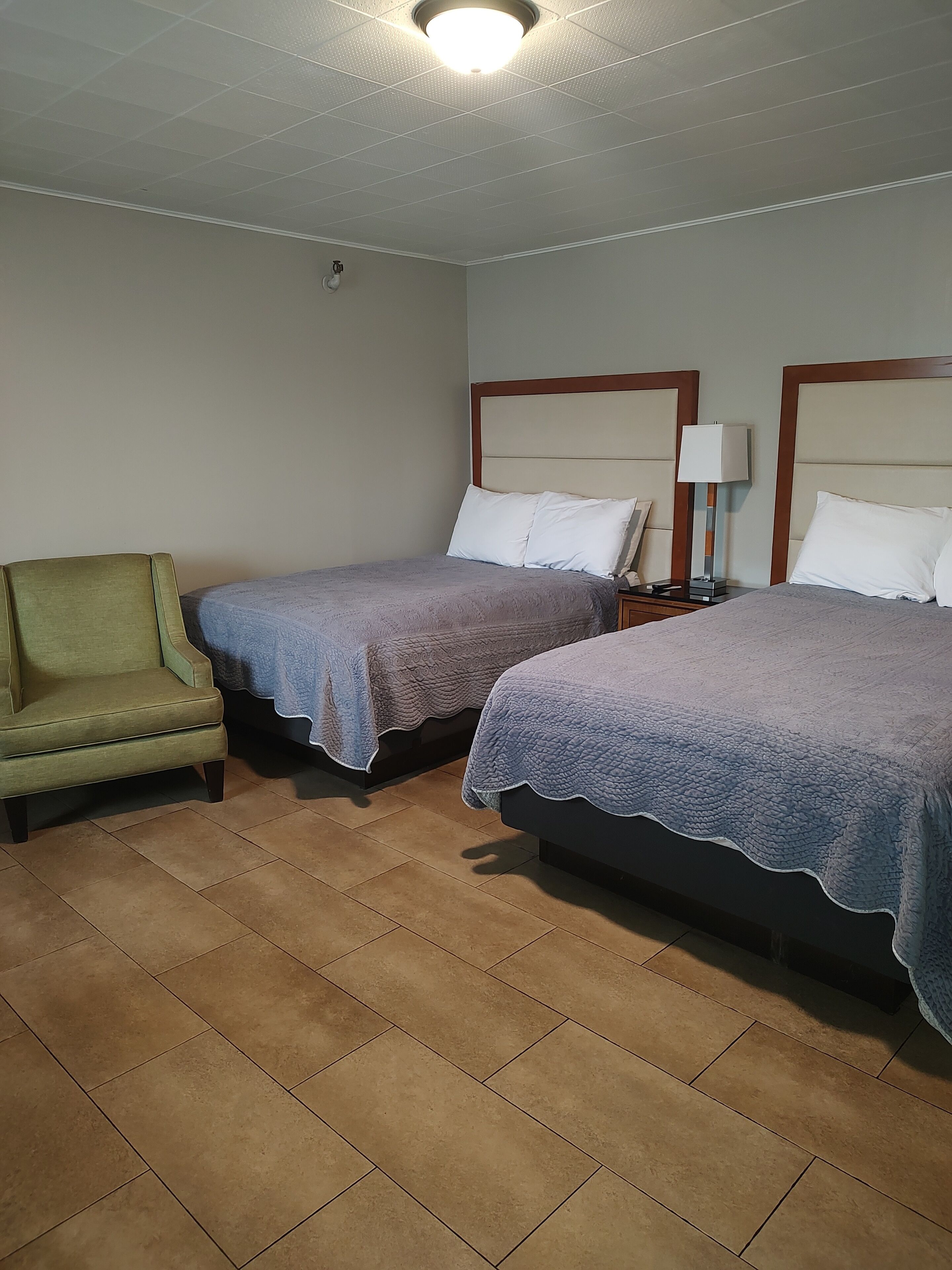 Et hotelværelse med to senge, en grøn lænestol og et natbord med en lampe.