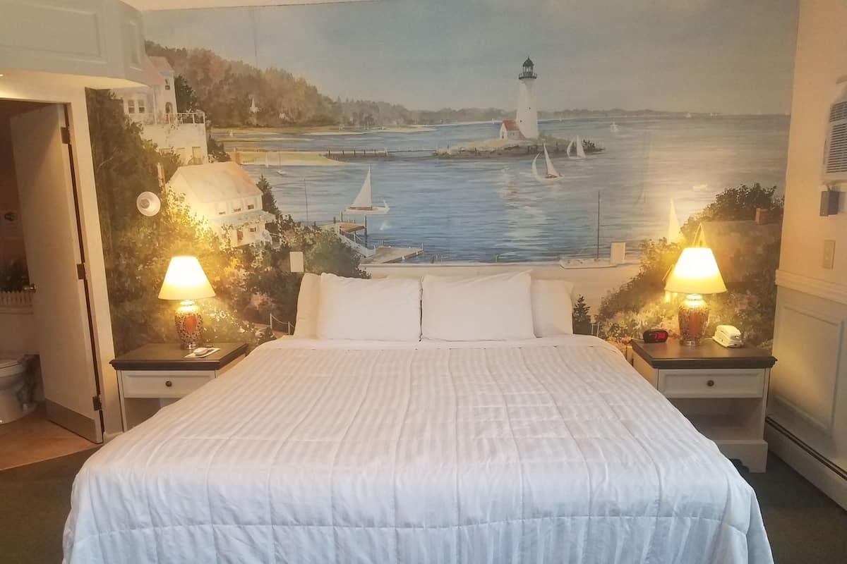 A bedroom with a bed, two bedside tables with lamps, a nightstand, and a wall mural of a coastal scene.
