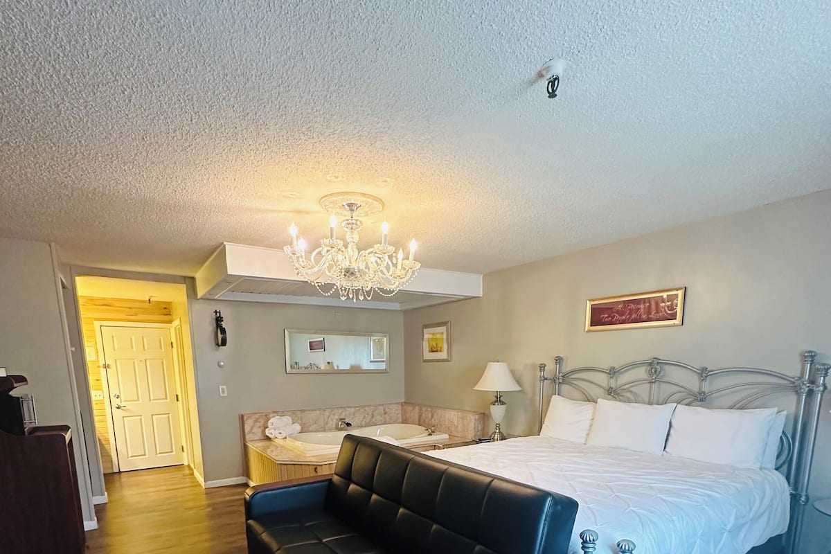 A bedroom with a bed, a black leather sofa, a chandelier, a bedside table, and a mirror.