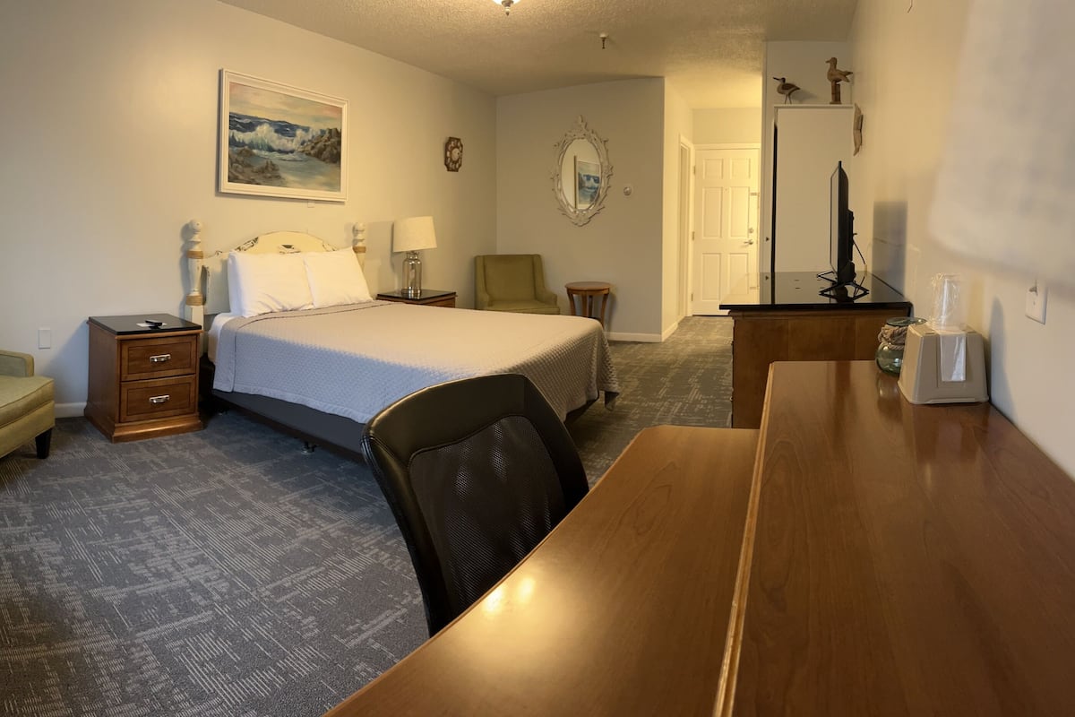 A hotel room with a bed, a desk, a chair, a nightstand, a lamp, a small table, and a mirror.
