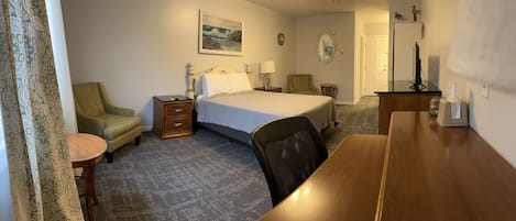 A hotel room with a bed, a desk, a chair, a nightstand, a lamp, a small table, and a mirror.