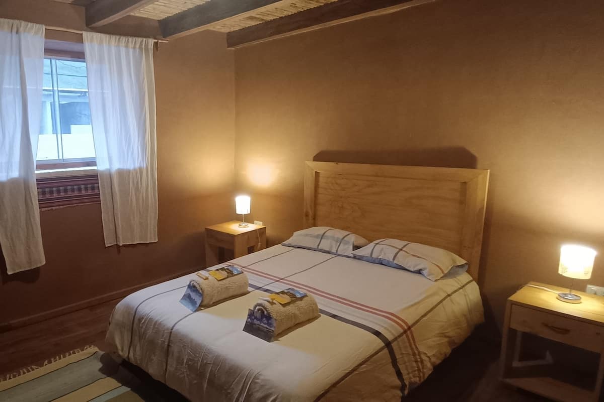 A hotel room with a wooden headboard, two bedside lamps, a window with white curtains, and a bed with towels.