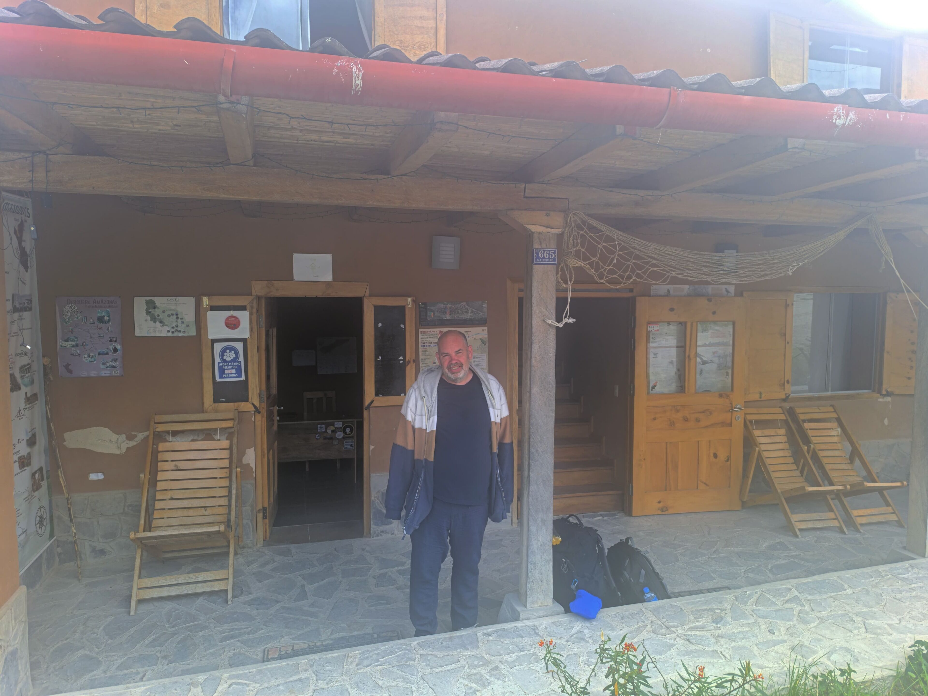 A man standing in front of a building with a wooden facade, a stone floor, and a red roof. There are wooden benches outside, a hammock, and a sign with the number 665.