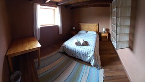 A small, cozy bedroom with a wooden ceiling, a single bed with a patterned bedspread, a small wooden table, and a window with curtains. - Muya (Tingo)