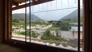A window with a view of a mountainous landscape, a stone wall, and some greenery. - Muya (Tingo)