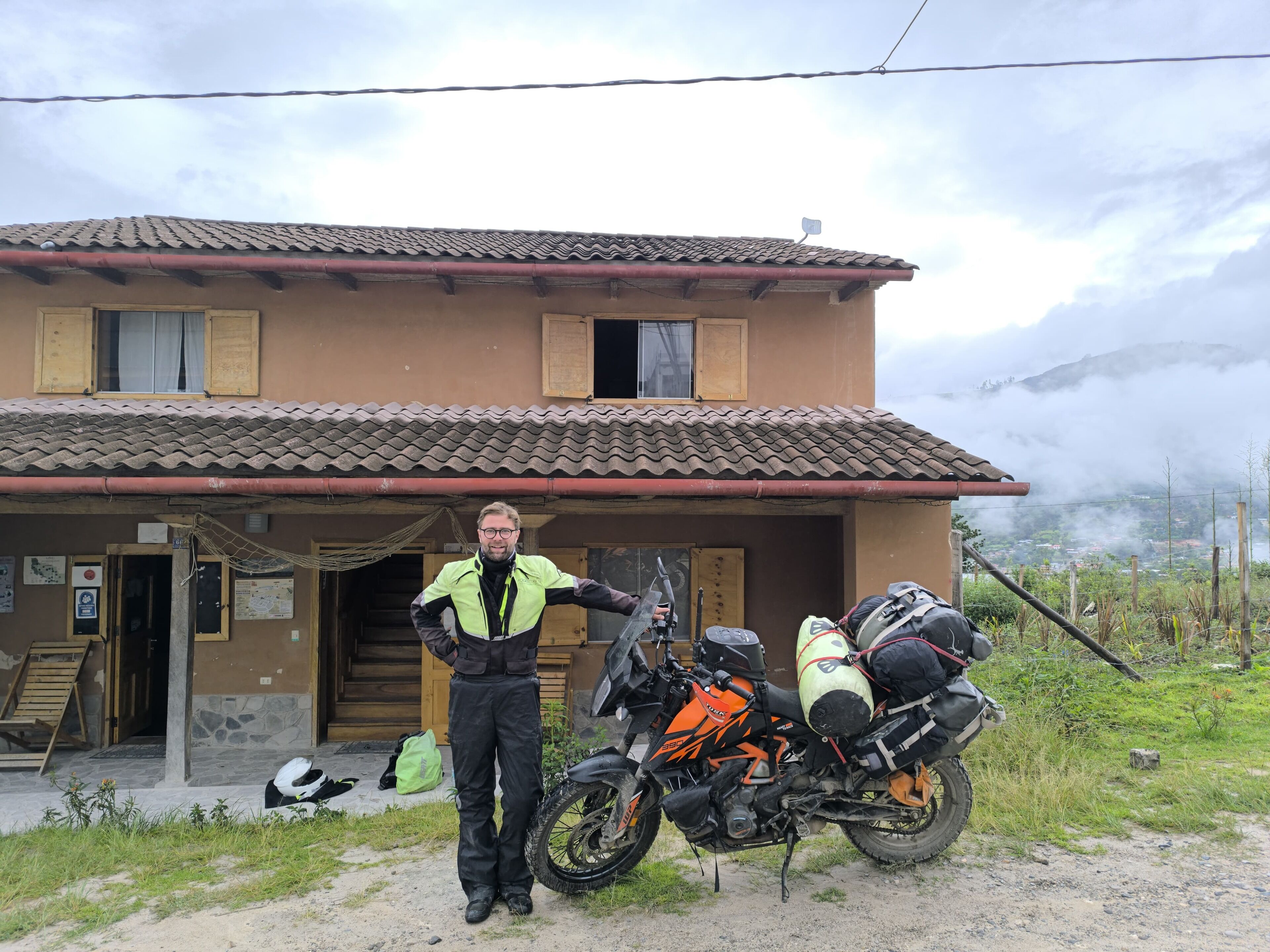 A person standing next to a motorcycle parked in front of a house with a tiled roof and a staircase leading to the entrance.