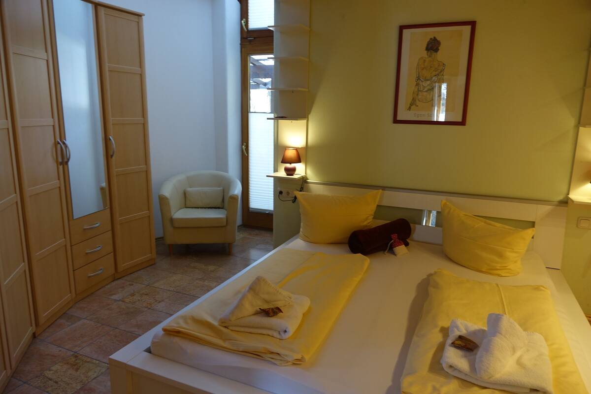 A hotel room with a bed, a chair, a wardrobe, and a nightstand with a lamp.