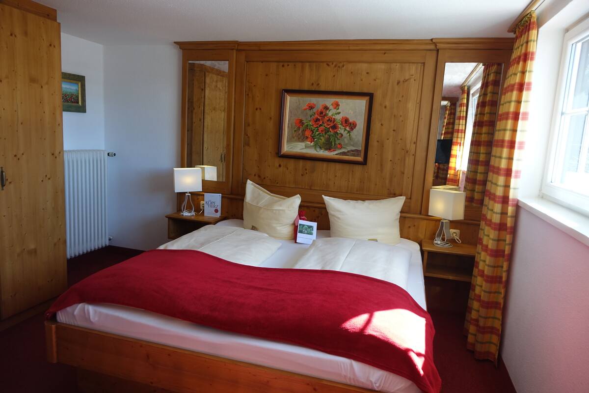 A wooden-paneled bedroom with a large bed, two bedside tables with lamps, a framed painting, and a window with curtains.