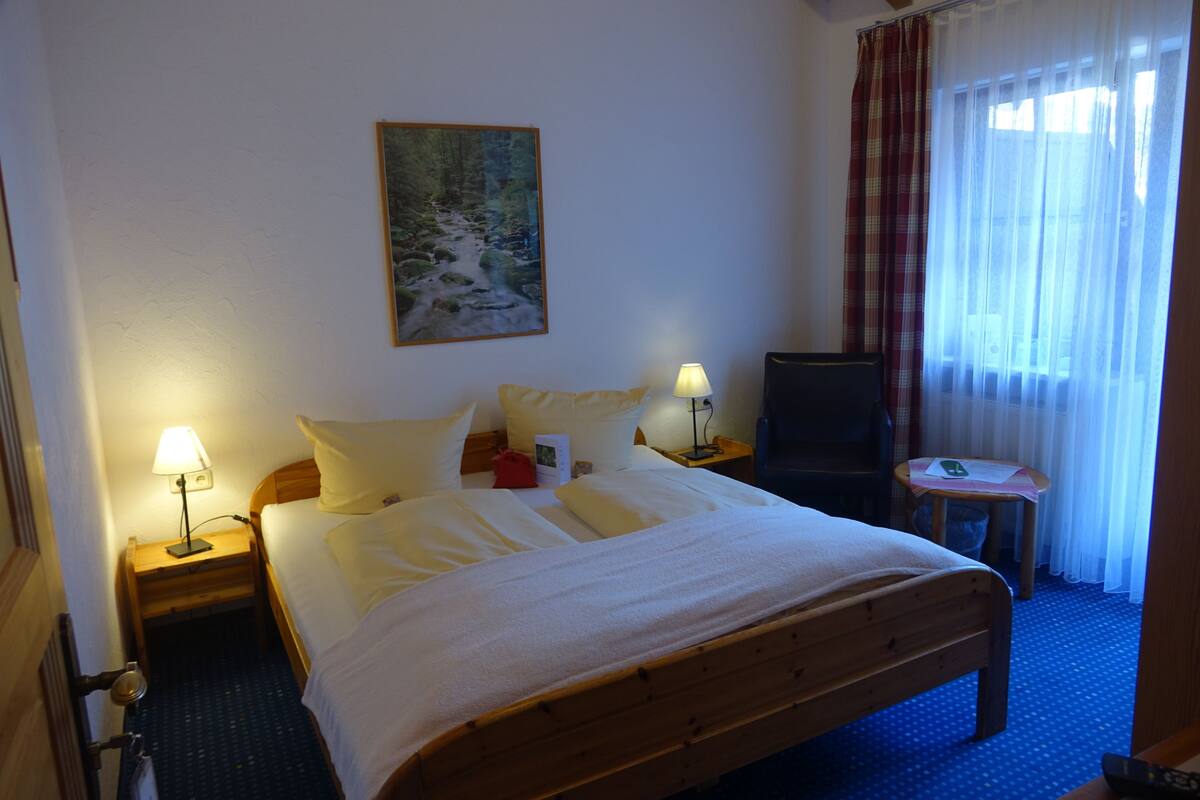 A hotel room with a bed, two bedside tables with lamps, a TV, a small table with a remote, and a painting on the wall.