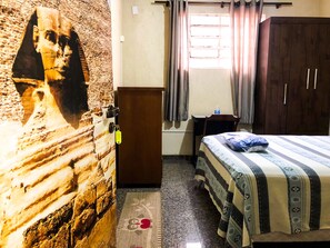 Una habitación con cama, armario, una mesita, un cuadro en la pared y una ventana con cortinas.