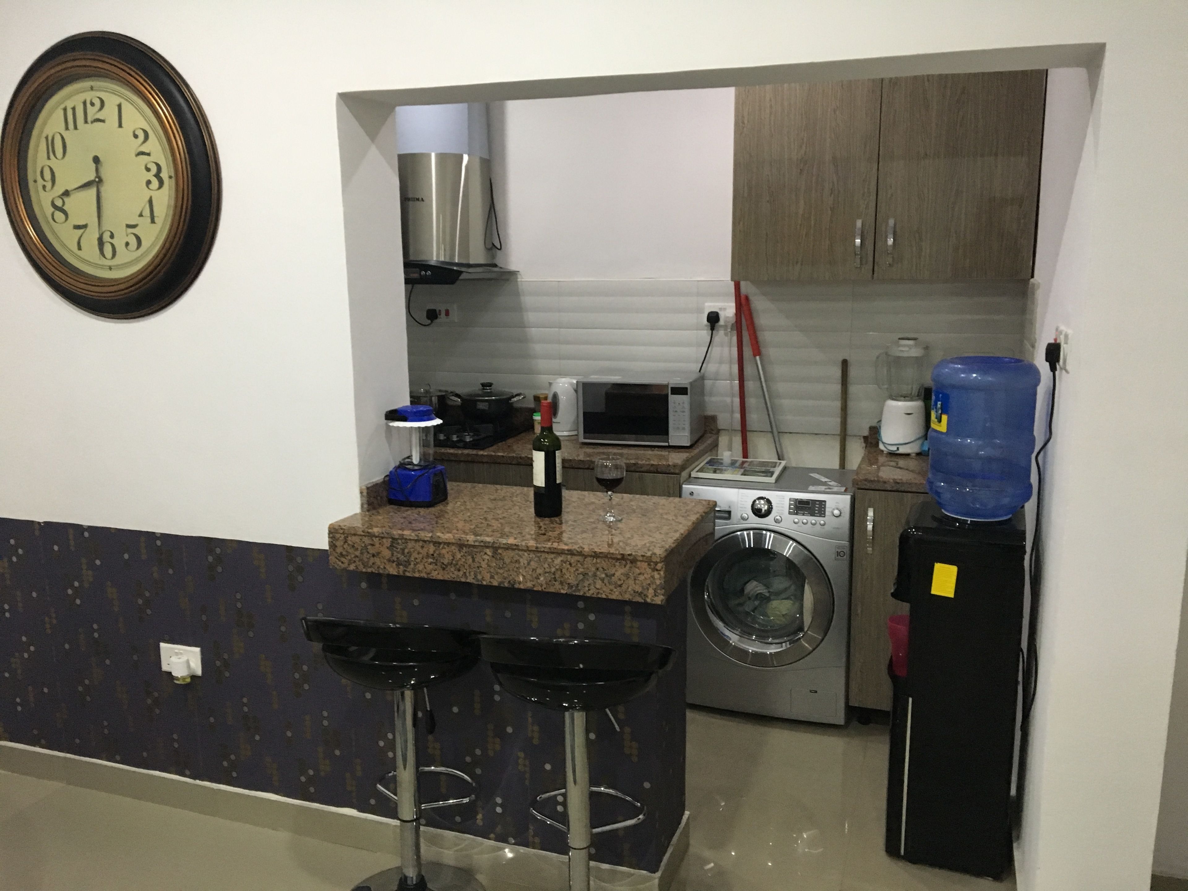 A compact kitchen with a washing machine, a microwave, a coffee maker, and a water dispenser. There are two bar stools and a clock on the wall.