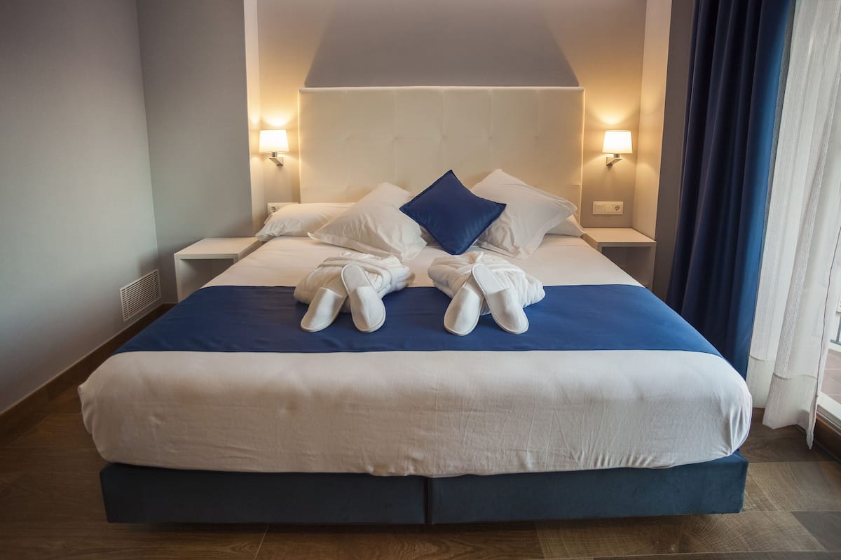 Un lit bien fait, recouvert d’une couvre-lit bleue et blanche, avec deux oreillers blancs et un oreiller décoratif bleu. On y trouve deux tables de chevet avec des lampes, ainsi qu’une fenêtre avec des rideaux.