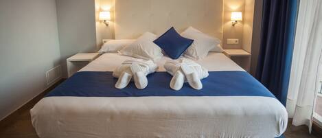 A neatly made bed with a blue and white bedspread, two white pillows, and a blue accent pillow. There are two bedside tables with lamps, and a window with curtains.