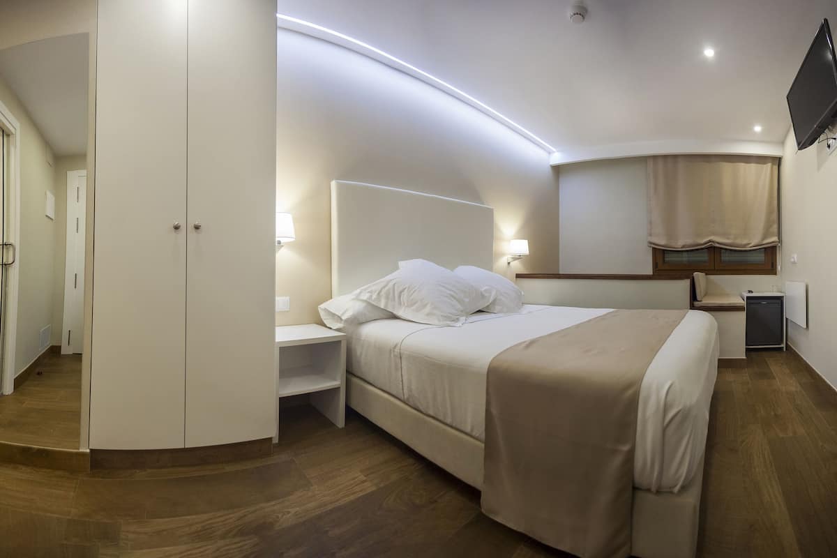 Une chambre d’hôtel moderne équipée d’un lit, d’une table de chevet et d’un petit bureau avec une lampe. On aperçoit une douche avec une porte en verre et des toilettes à l’arrière-plan.