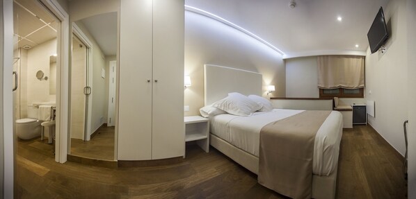 A modern hotel room with a bed, bedside table, and a small desk with a lamp. There is a shower area with a glass door and a toilet visible in the background.