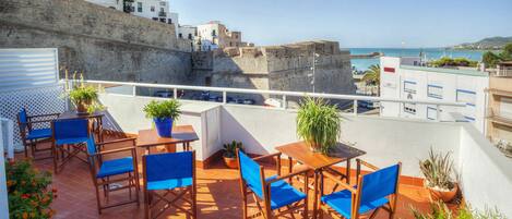 A terrace with wooden tables and blue chairs, potted plants, and a view of the sea and buildings.