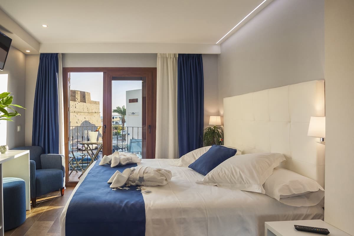 Une chambre d’hôtel équipée d’un lit, d’un bureau, d’une télévision et d’un balcon offrant une vue sur un bâtiment historique.