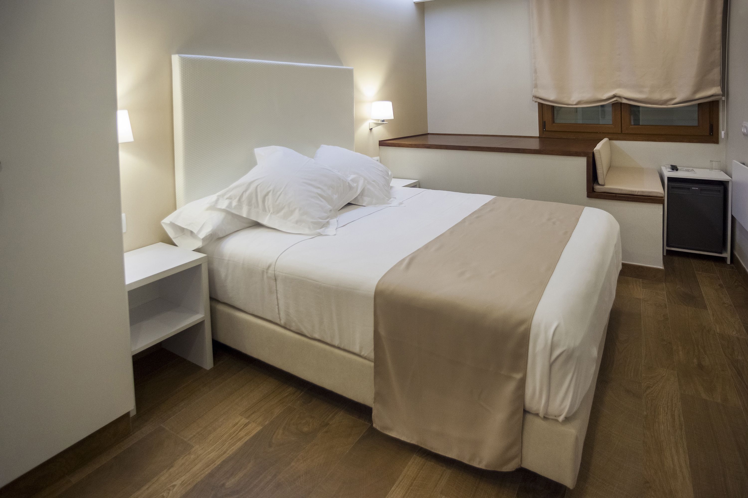 Een netjes ingerichte hotelkamer met een bed, nachtkastjes, een klein bureau, een lamp, een raam met gordijnen en een houten vloer.