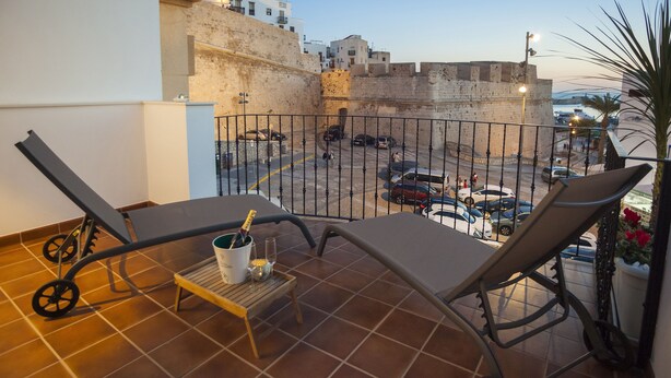 Un balcon avec deux fauteuils, une petite table sur laquelle se trouve une boisson, et une vue sur une rue animée avec des voitures et des immeubles.