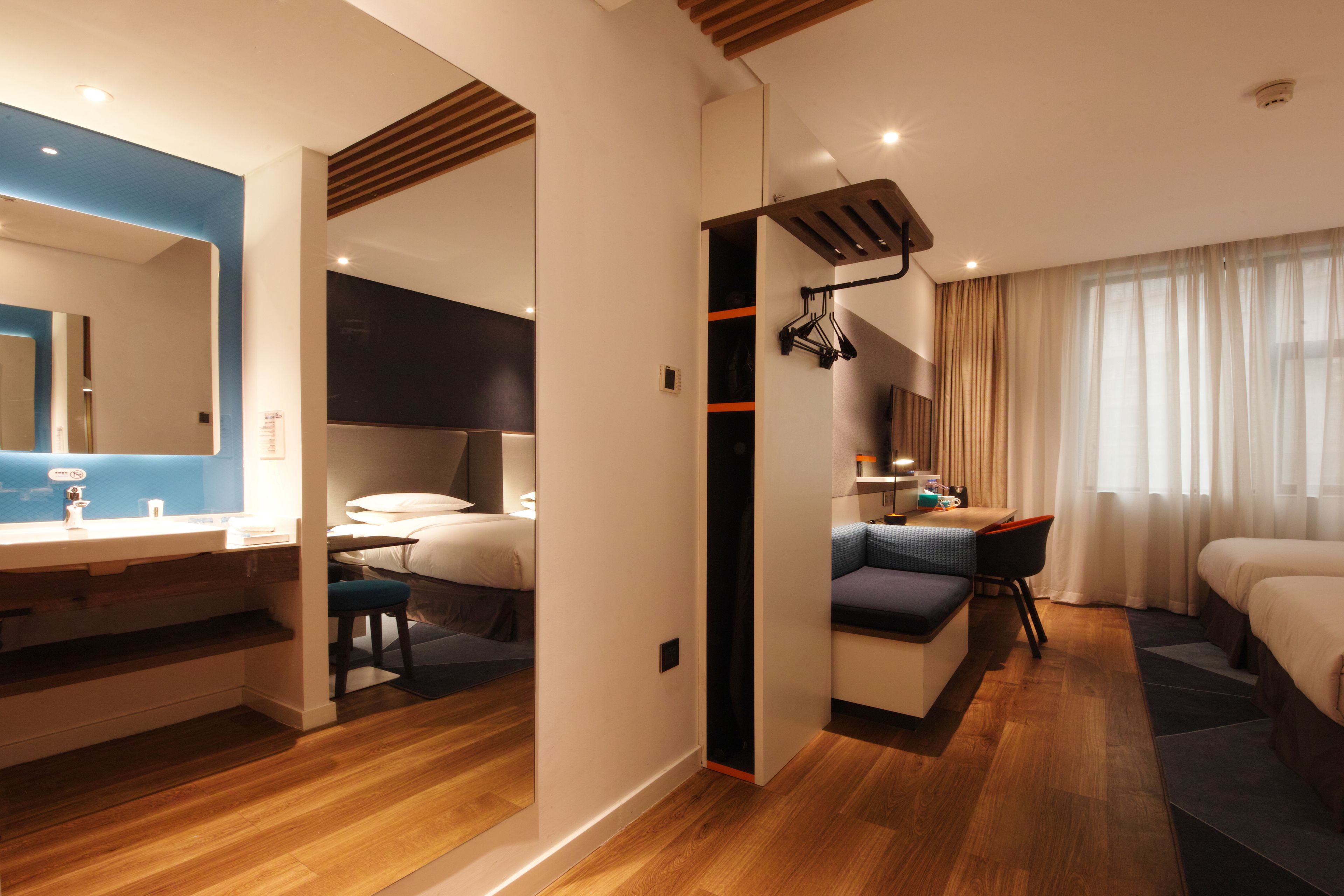 A modern hotel room with a wooden floor, a large mirror, a desk with a chair, a sofa, and a bed.