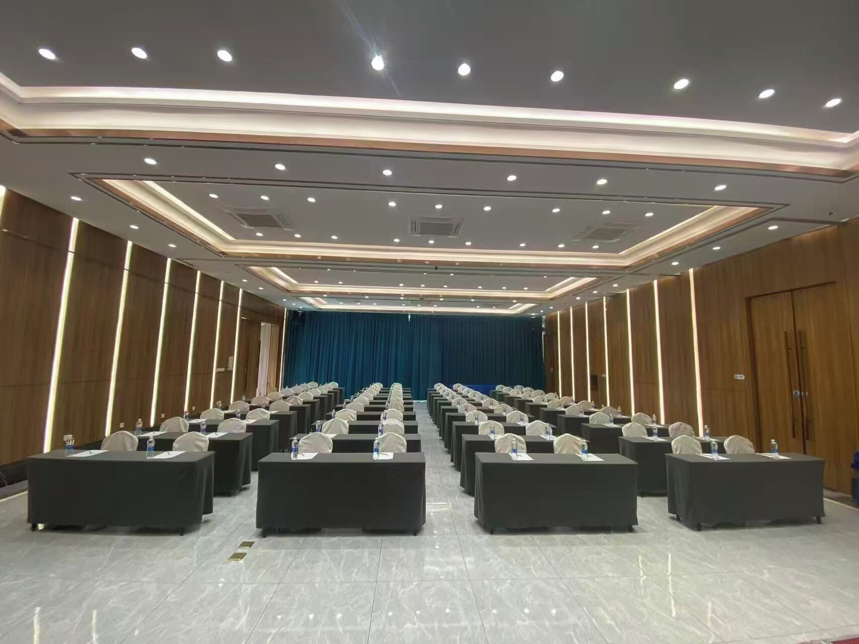 A conference room with rows of tables and chairs, a stage with a blue curtain, and a modern ceiling design.
