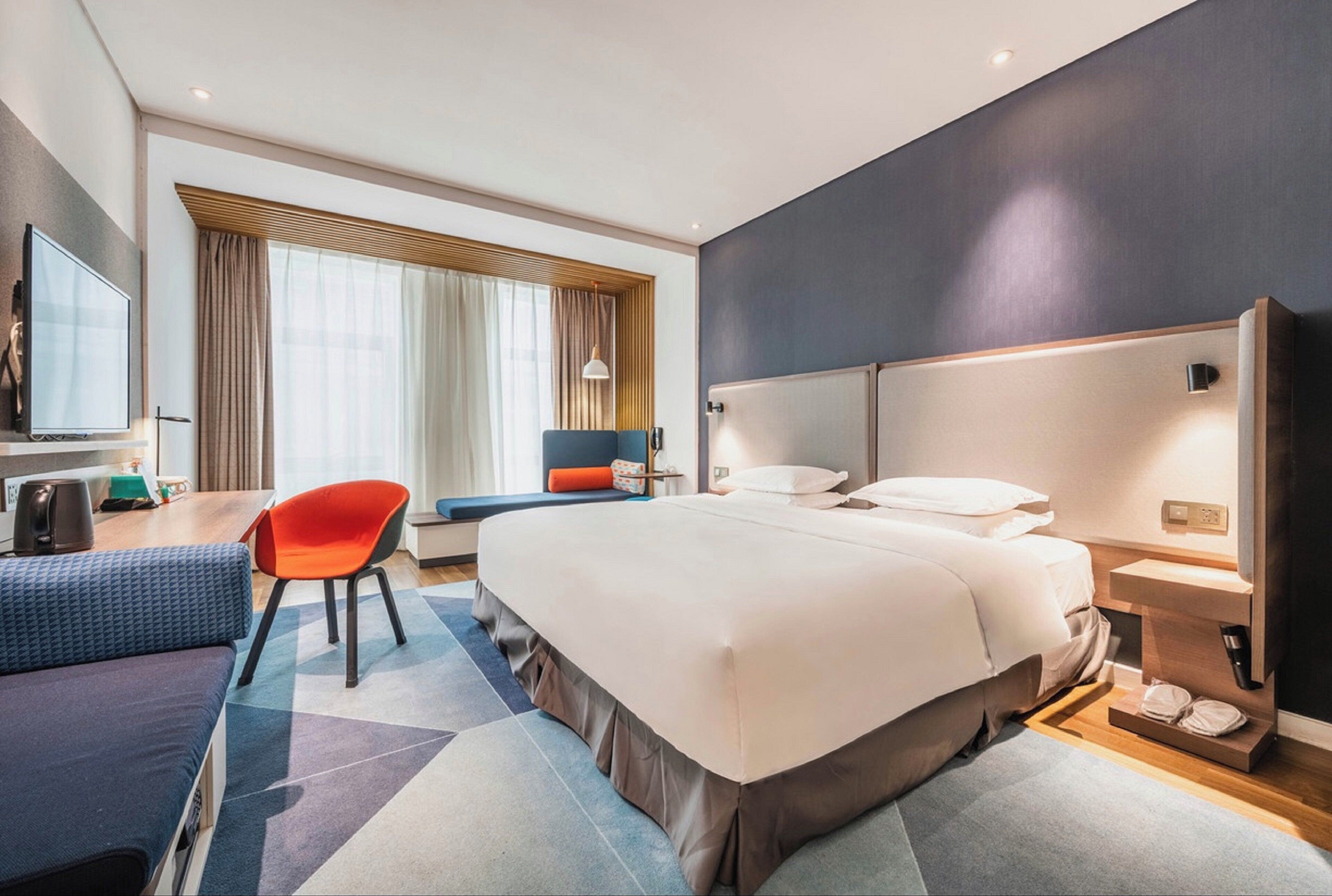 A modern hotel room with a large bed, a desk with a chair, a television, and a window with curtains.