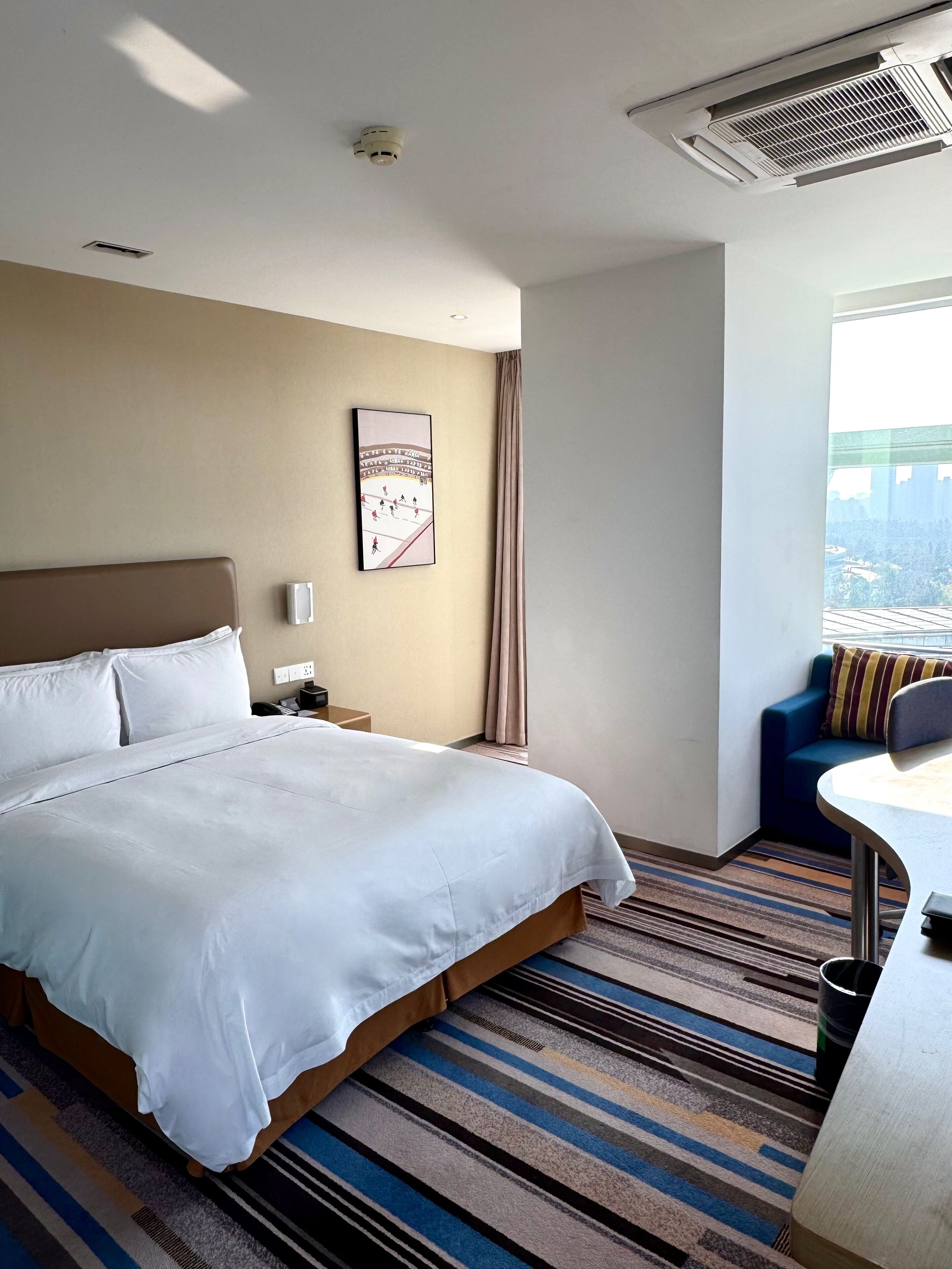 A hotel room with a large bed, a desk, a chair, a window with a city view, and a ceiling vent.