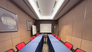 Un salón de conferencias con una larga mesa, sillas rojas, una pizarra y una pantalla de proyector.