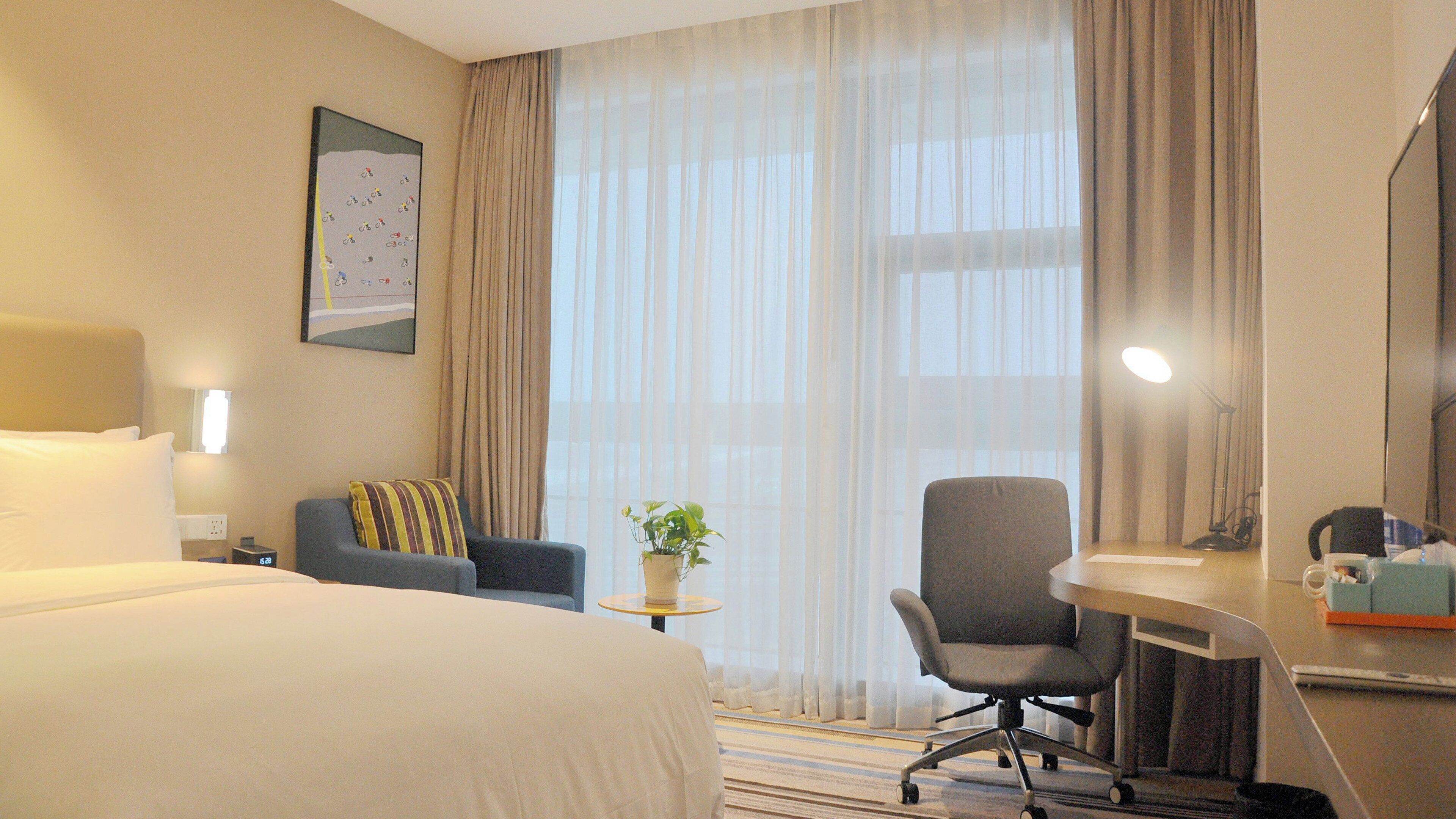 A hotel room with a large bed, a desk with a chair, a small table with a plant, and a window with curtains.