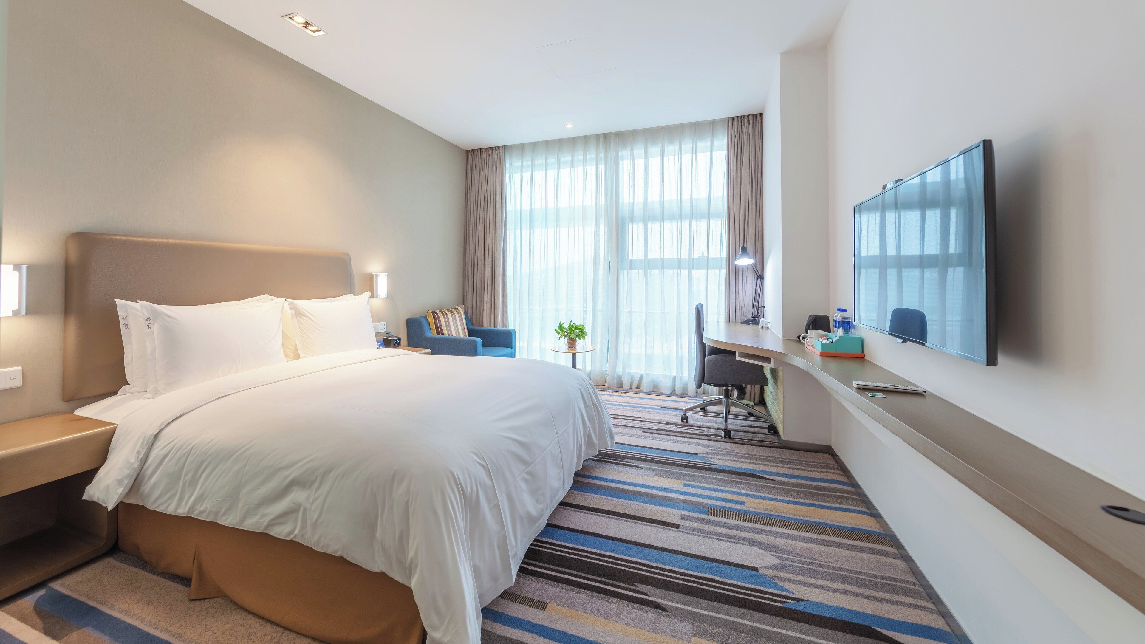 Habitación de hotel con cama, escritorio, silla, televisor y ventana con cortinas.