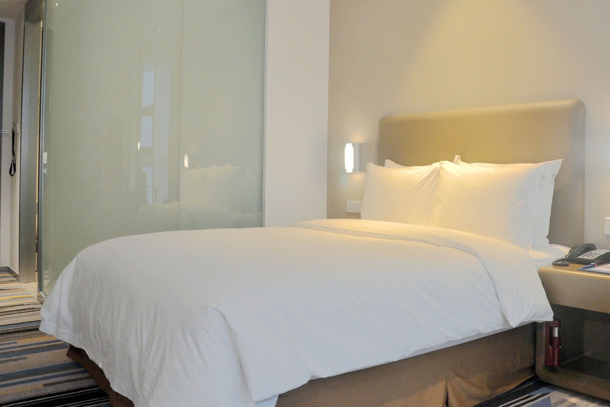 Una habitación de hotel con una cama, mesita de noche, lámpara y una planta.