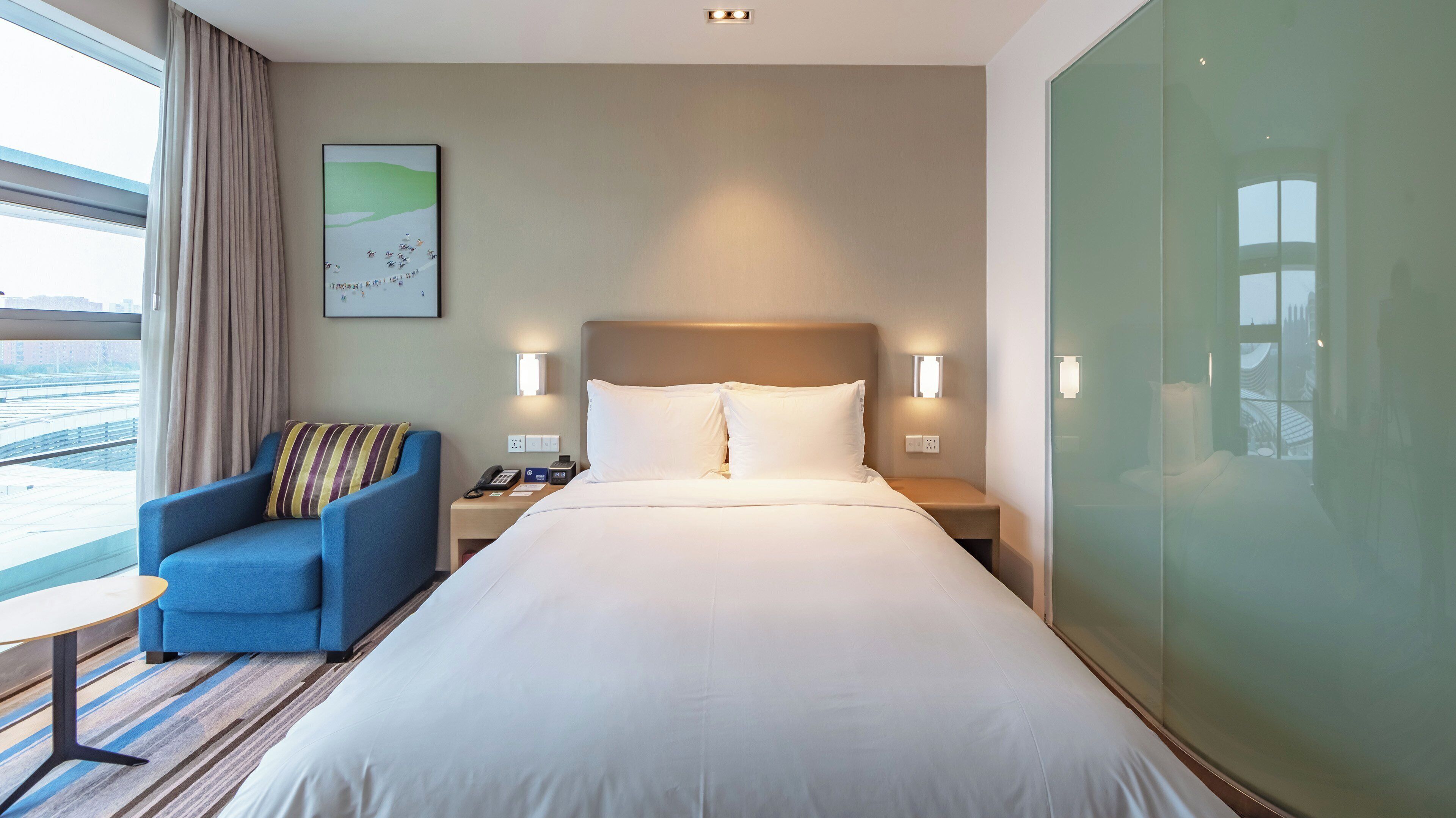 A hotel room with a large bed, a blue armchair, a small table, and a view of the outside.