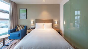 A hotel room with a large bed, a blue armchair, a small table, and a view of the city through the window.