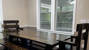Un espace repas avec une table et des bancs en bois, de grandes fenêtres équipées de stores et un mur blanc.