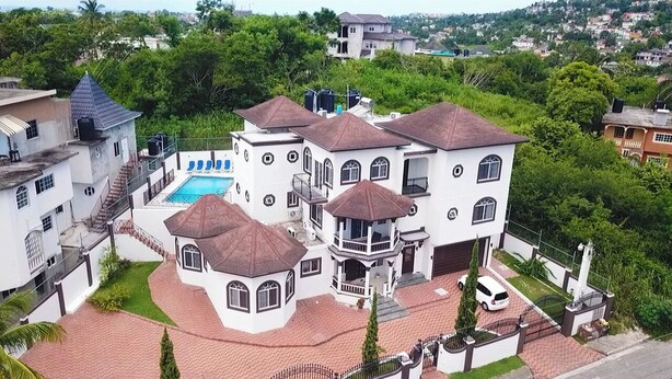 A large, multi-story house with a swimming pool, surrounded by lush greenery and other residential buildings.