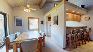 A dining area with a long table and chairs, a ceiling fan, and a kitchen with wooden cabinets and a clock on the wall.