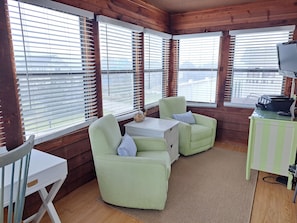 Interior acogedor de una cabaña con paredes de madera, un escritorio blanco, dos sillones verdes, una mesita blanca y un televisor sobre un mueble verde.