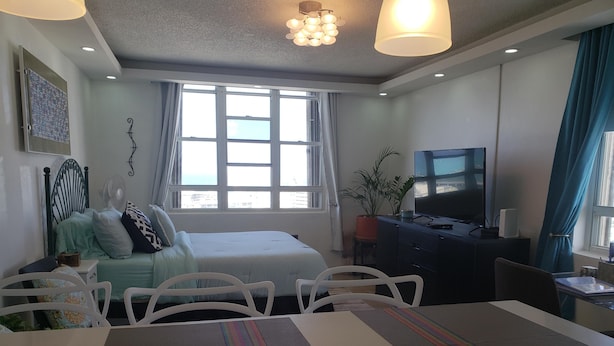 A modern hotel room with a bed, a desk with a computer, a TV, and a large window with curtains. - Apex living in Puerto Rico's capital  (Santurce)