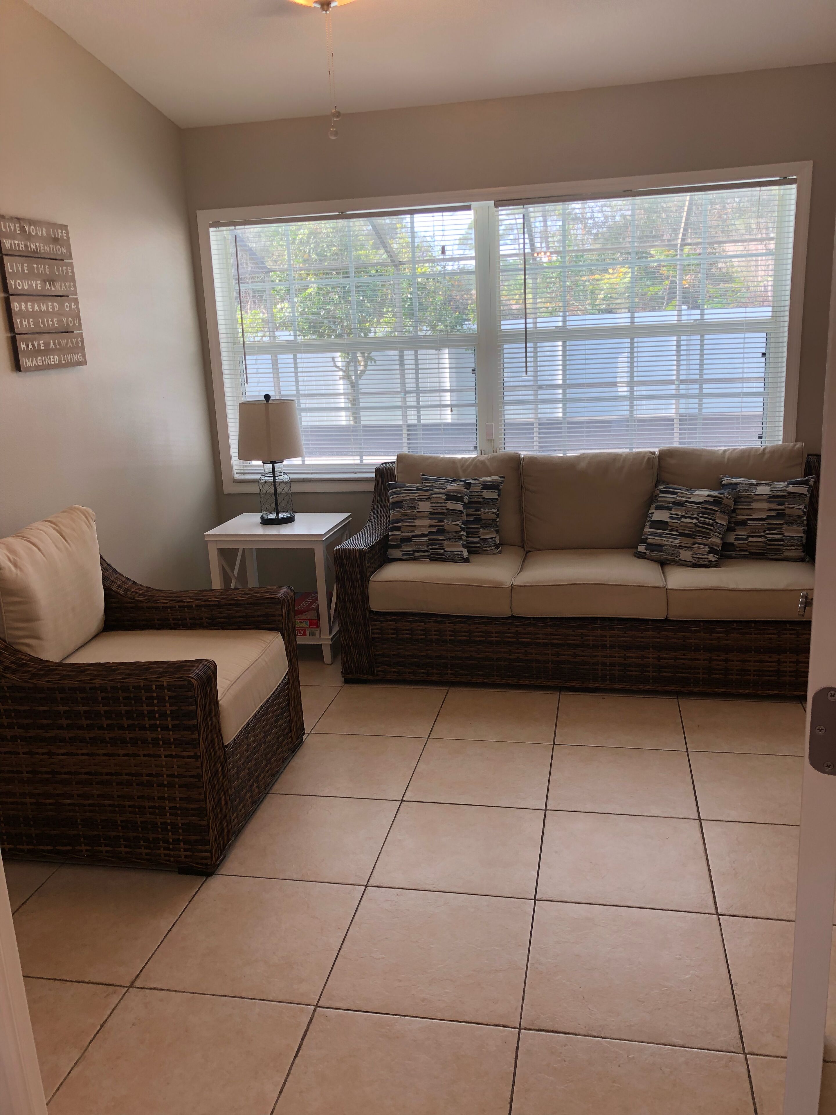 A living room with a beige sofa, a wicker chair, a small table with a lamp, and a window with blinds.