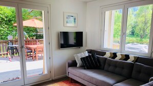 Een woonkamer met een grijze bank, een flatscreen televisie en uitzicht op de natuur door grote ramen.