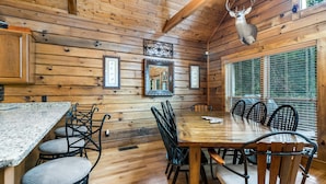 Interior de uma cabana de madeira com mesa de jantar, cadeiras, quadros e um chifre de veado montado na parede.