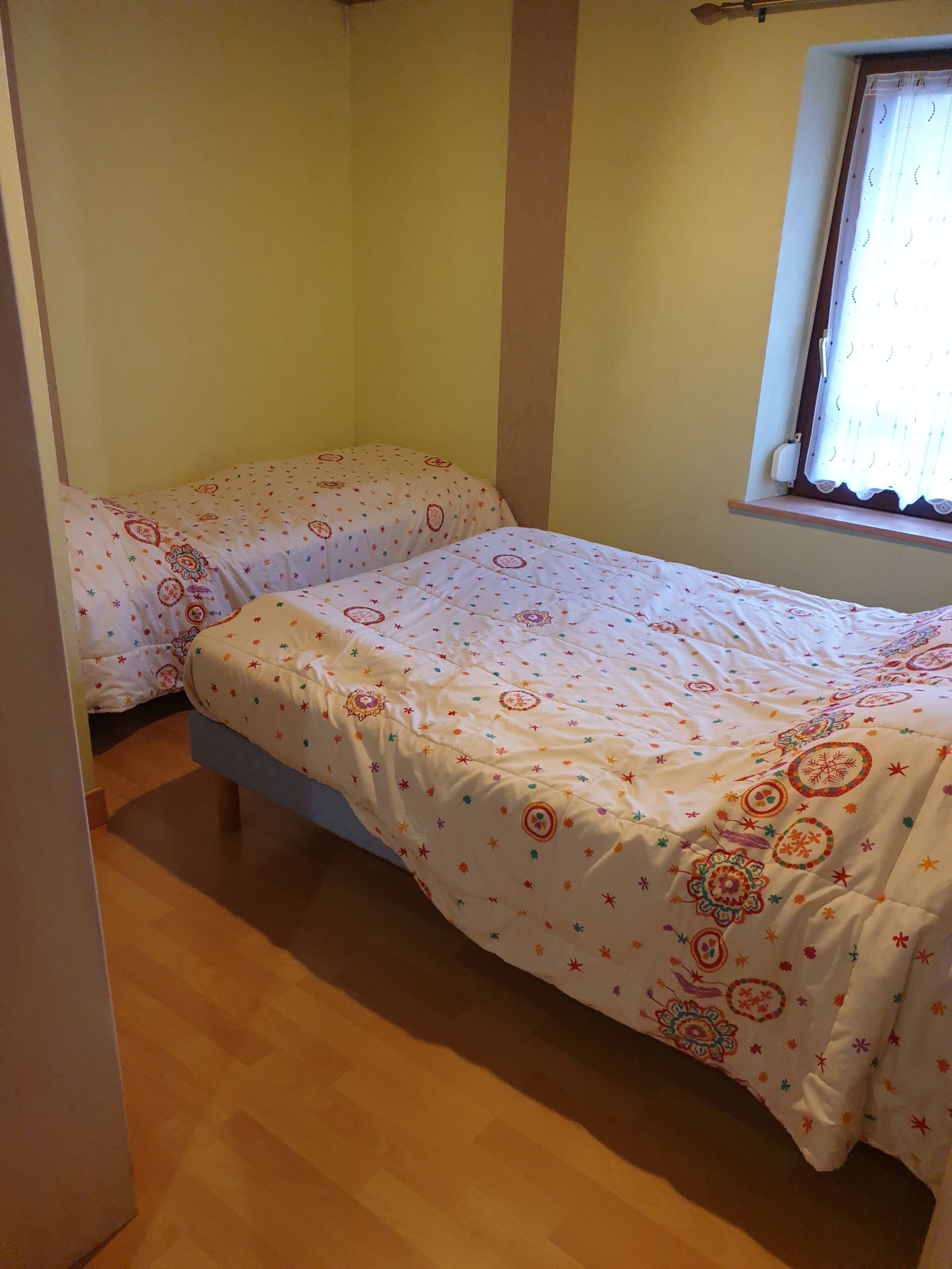 A small, single-bed room with a wooden floor, a window with a curtain, and a bed with a patterned bedspread.