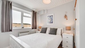A bedroom with a large bed, bedside tables, a window with curtains, a radiator, a wall-mounted picture, and a hanging plant.