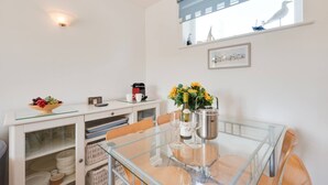 A dining area with a glass table, chairs, a coffee machine, a vase with flowers, and a window with a view of a building.